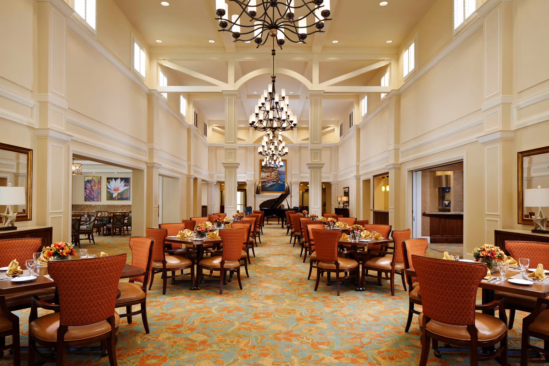 A spacious and elegant dining room with high ceilings and large chandeliers. The room features multiple round tables with orange upholstered chairs, each table set with plates, glasses, napkins, and floral centerpieces. At the far end of the room, there is a grand piano and a large painting above a fireplace. The walls are painted in a light cream color with decorative molding, and there are large mirrors on the side walls.
