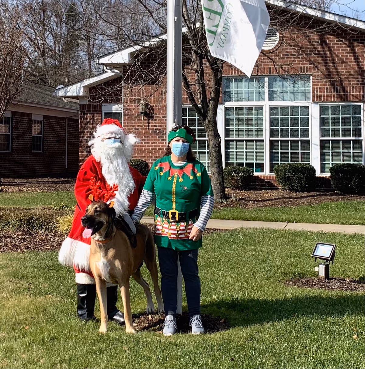 A person dressed as Santa Claus and another person dressed as an elf, both wearing face masks, stand outside on a grassy area in front of a brick building. A large dog with reindeer antlers stands in front of them. A flagpole with a white flag is visible behind them.