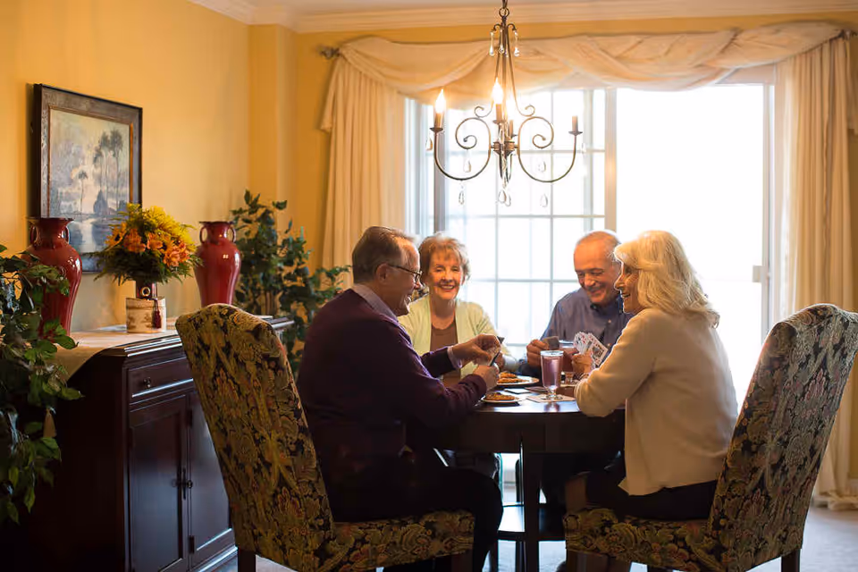 Four elderly people sitting around a dining table playing cards in a warmly lit room with large windows covered by curtains. The room has floral upholstered chairs, a chandelier, a sideboard with red vases, a flower arrangement, and a framed painting on the wall.