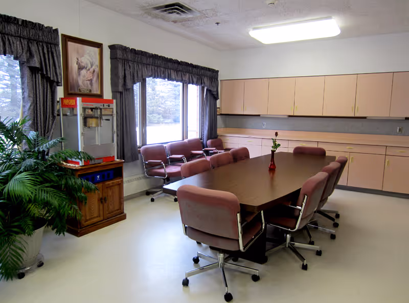 Conference-style dining room with a long table surrounded by wheeled upholstered chairs, cabinets along the wall, and a popcorn machine by the window.