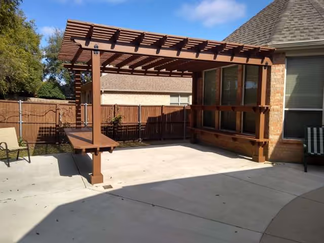 A wooden pergola extends over a concrete patio beside a brick building in a fenced yard.