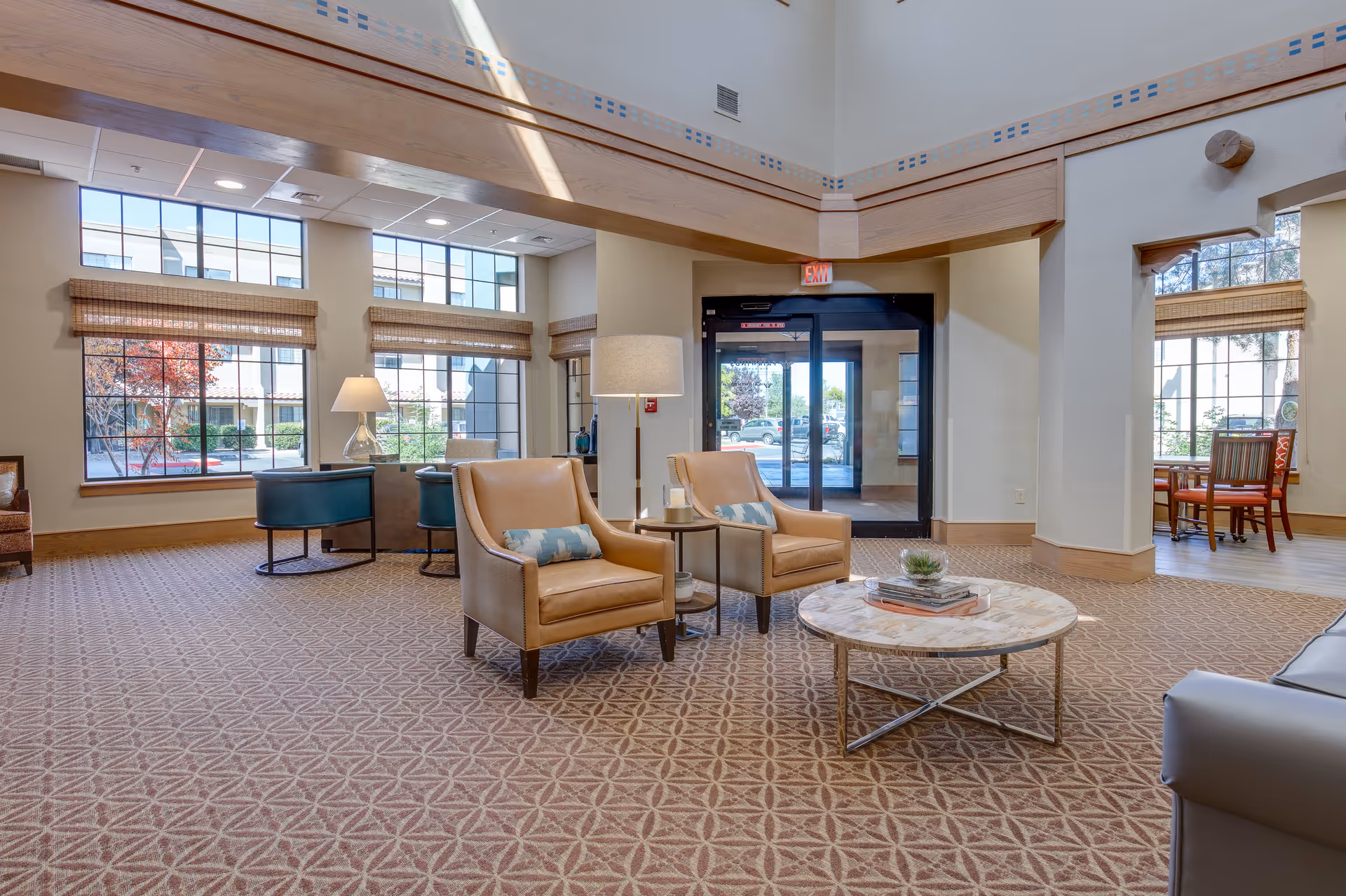 A spacious and well-lit living room area with large windows allowing natural light to fill the space. The room features two tan leather armchairs with decorative pillows, a round marble coffee table with books and a small plant, a floor lamp, and additional seating near the windows. The entrance door with glass panels is visible in the background.