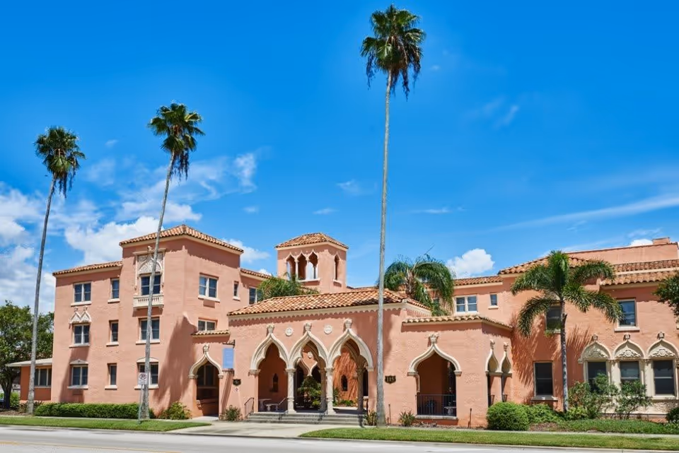 Exterior view of a large pink stucco building with Spanish-style architecture, featuring arched doorways and windows, red tile roof, and tall palm trees in front under a clear blue sky.