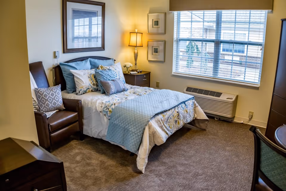 A cozy bedroom with a neatly made bed featuring blue and white bedding and multiple pillows. Next to the bed is a wooden nightstand with a lamp and decorative items. A brown leather armchair with a patterned cushion is positioned beside the bed. The room has a large window with blinds, a wall-mounted air conditioning unit below the window, and framed artwork on the walls. The floor is carpeted in a neutral tone.