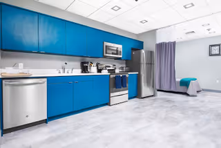 A modern kitchenette with bright blue cabinets, stainless steel appliances including a dishwasher, stove, microwave, and refrigerator. The room has a light gray floor and ceiling tiles with recessed lighting. In the background, there is a bed partially visible with a teal blanket and a purple privacy curtain.