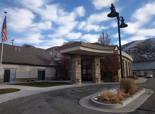 Exterior view of a single-story assisted living facility with stone pillars at the entrance, a curved driveway, a flagpole with an American flag, and mountains in the background under a partly cloudy sky.