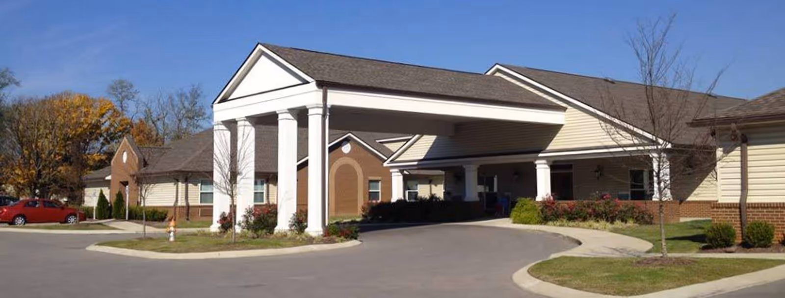 Front exterior view of Grace Manor Assisted Living building with a covered entrance supported by white columns, surrounded by a paved driveway, landscaped grass, and trees with autumn foliage.