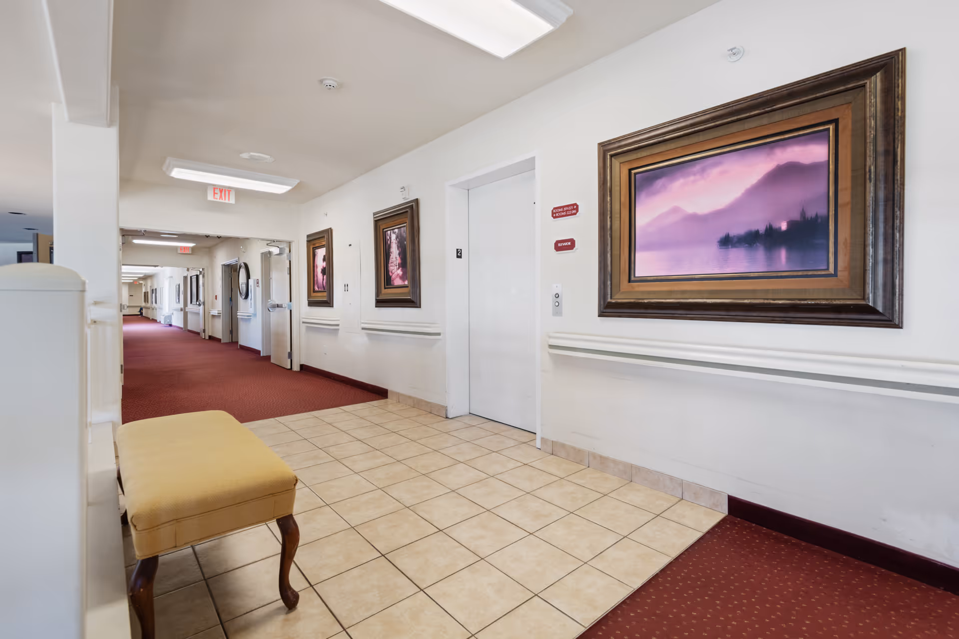 Interior hallway of a senior living facility with beige tiled floor near an elevator and red carpeted corridor. There is a yellow cushioned bench on the left, framed landscape paintings with purple and pink hues on the walls, and exit signs visible in the distance.