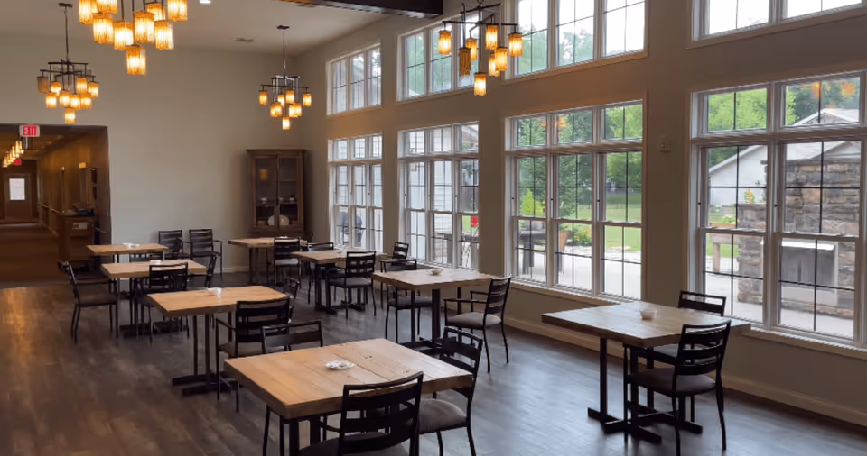 A dining area with multiple wooden tables and black chairs arranged neatly. Large windows on one side let in natural light and offer a view of greenery outside. Several pendant lights hang from the ceiling, illuminating the space. A hallway is visible in the background.
