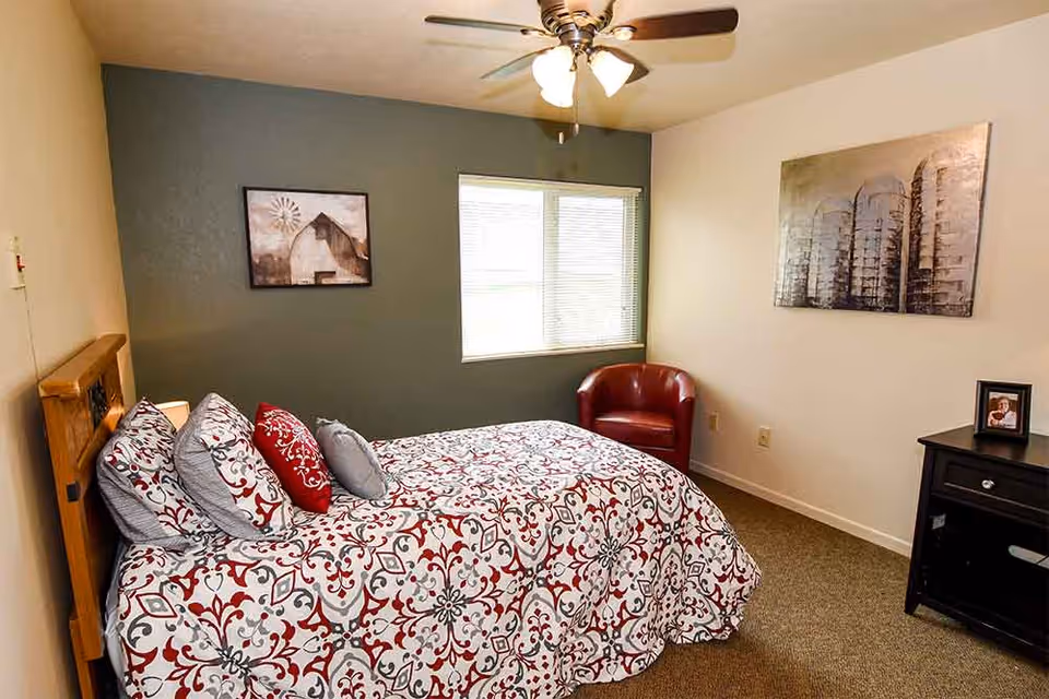 A bedroom with a patterned red-and-gray bedspread, several pillows, a red armchair, window, and wall art.