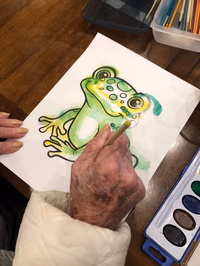 An elderly person painting a colorful frog illustration on a piece of paper using watercolors at a wooden table. Various painting supplies, including a watercolor palette and colored pencils, are visible nearby.
