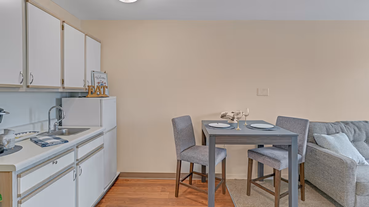 A small kitchen and dining area with white cabinets, a white refrigerator, a sink, and a countertop. Next to the kitchen is a gray dining table set with two gray upholstered chairs, two plates, and two wine glasses. A gray sofa with a light-colored pillow is partially visible on the right side.