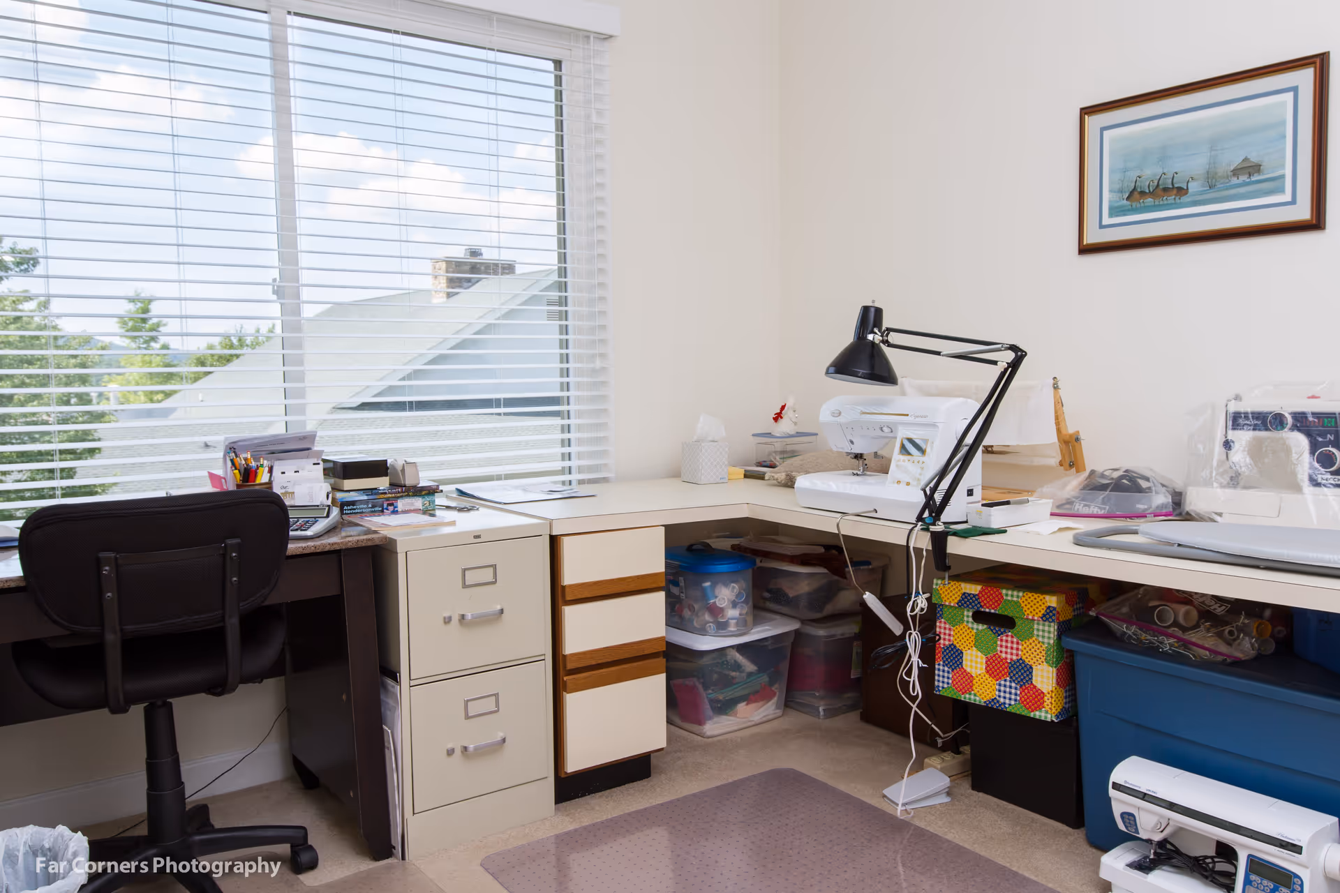 A bright craft room with a large window covered by white blinds. The room features a black office chair, a desk with a filing cabinet, and a corner workspace with a sewing machine, desk lamp, and various storage bins containing craft supplies. A framed picture of geese is hung on the wall.