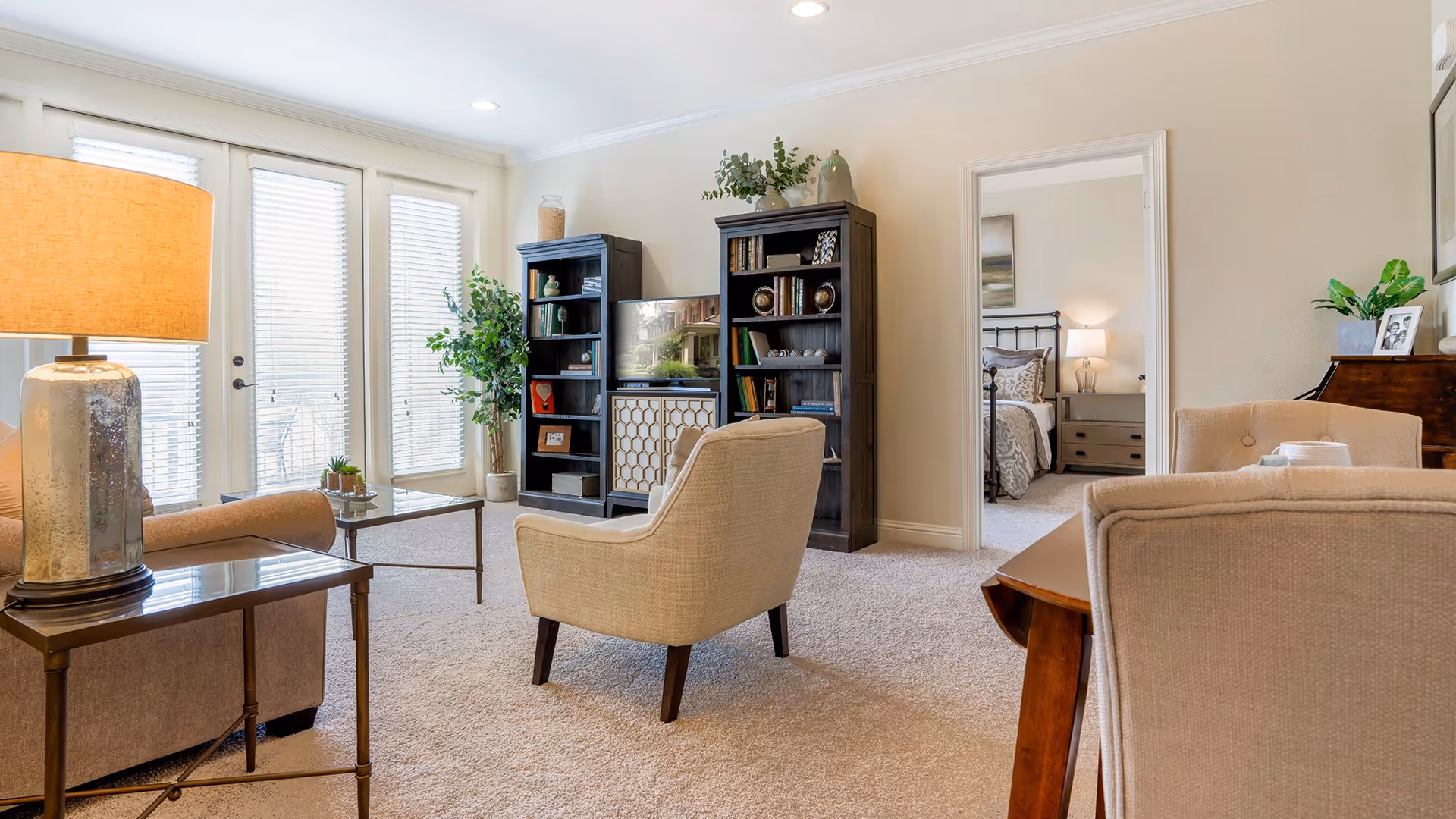 A bright living room with beige carpet and light-colored walls. The room features a beige armchair, a side table with a lamp, a coffee table, and two black bookshelves with decorative items and a TV. French doors with blinds lead outside, and a doorway reveals a bedroom with a bed and nightstand with a lamp.
