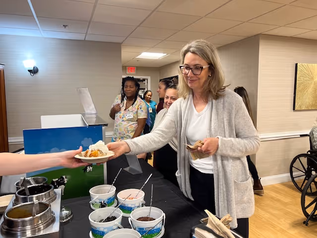 A woman wearing glasses and a light gray cardigan is receiving a plate of food from a server at a buffet table inside a facility. Several other people, including staff in colorful scrubs, are standing in line behind her. The room has beige walls, wood flooring, and a wheelchair is visible on the right side.
