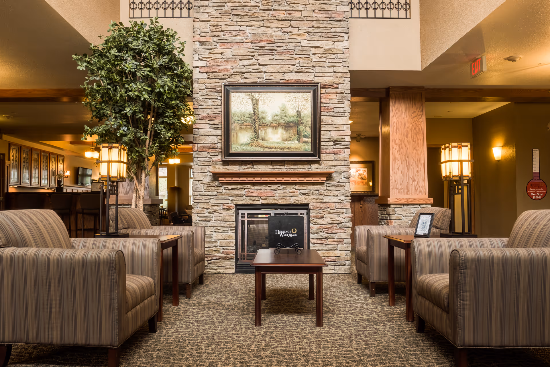 A cozy seating area in a senior living facility with four striped armchairs arranged around two wooden side tables and a central coffee table. The space features a stone fireplace with a framed landscape painting above it, a tall indoor plant, warm lighting from lamps, and a carpeted floor.