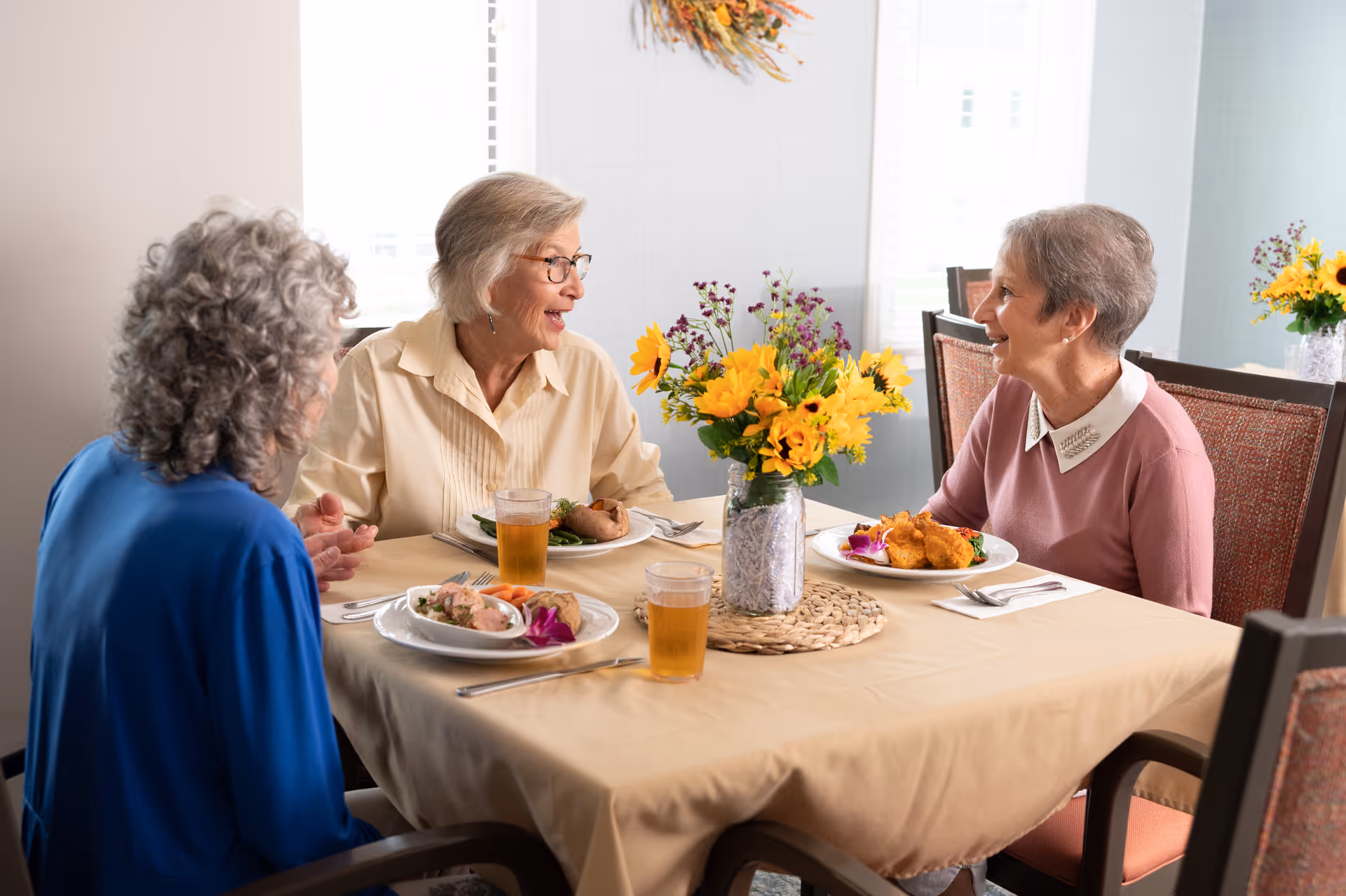 Three older women seated around a dining table with plates of food and a vase of yellow flowers, chatting.