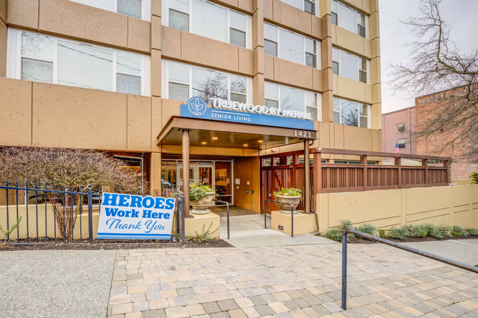 Entrance of the Truewood by Merrill senior living building with a covered doorway, potted plants, and a 'Heroes Work Here Thank You' sign.