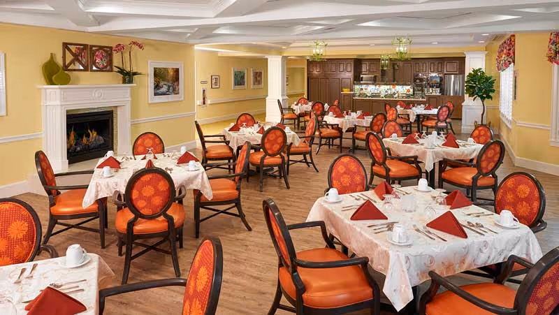 Bright dining room with multiple tables set with white tablecloths, red napkins and orange upholstered chairs, a fireplace on the left and a serving area in the back.