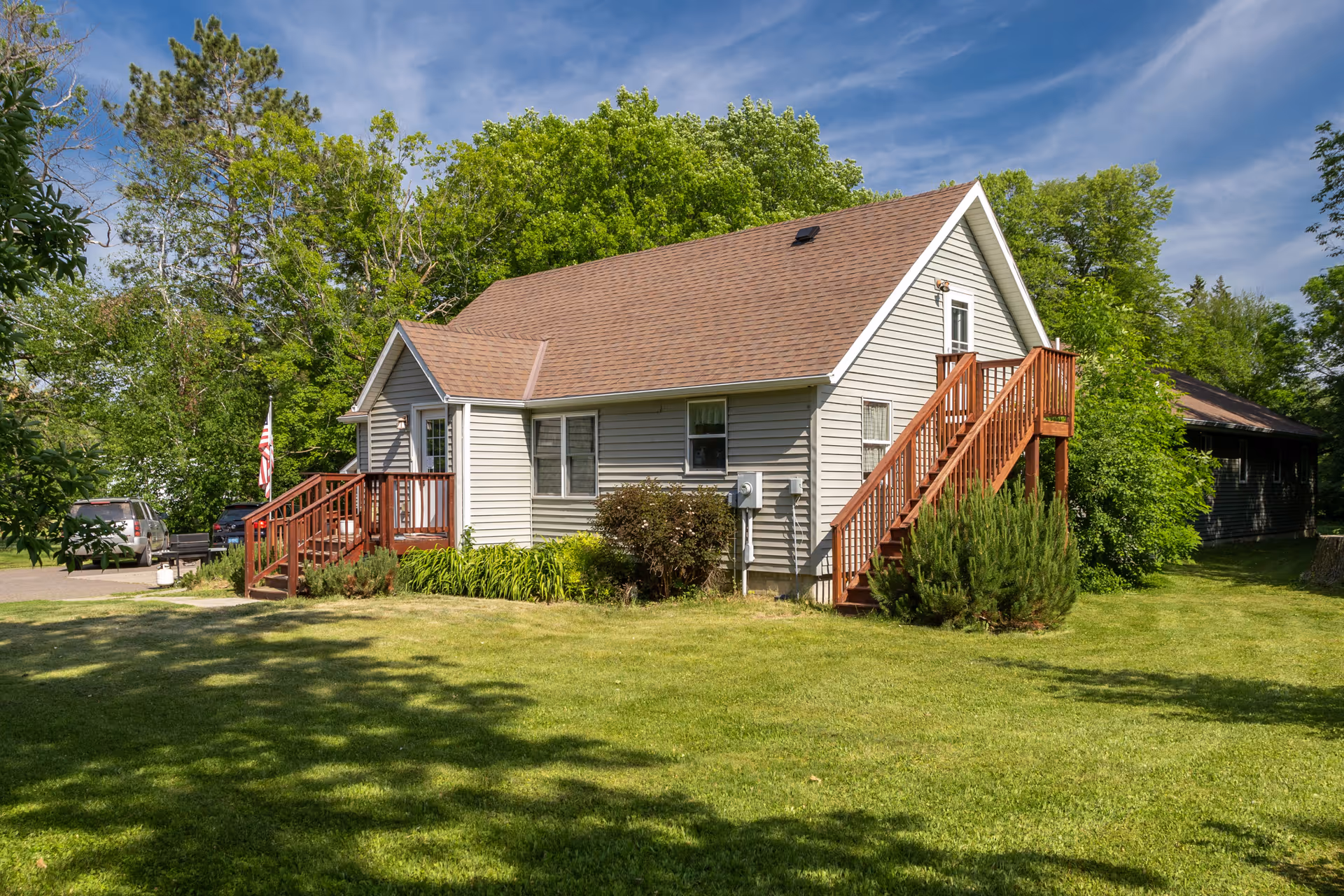 A small, single-story house with beige siding and a brown shingled roof surrounded by green grass and trees under a partly cloudy blue sky. The house has two sets of wooden stairs, one at the front entrance and one leading to a door on the upper level at the side. There are shrubs and plants around the base of the house, and a few parked cars are visible in the background.