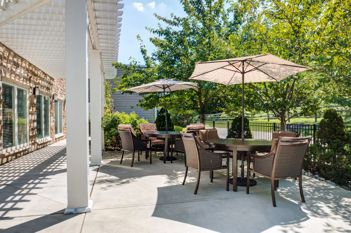 Outdoor patio area with round tables and chairs under large umbrellas, surrounded by greenery and trees, adjacent to a building with stone walls and windows.