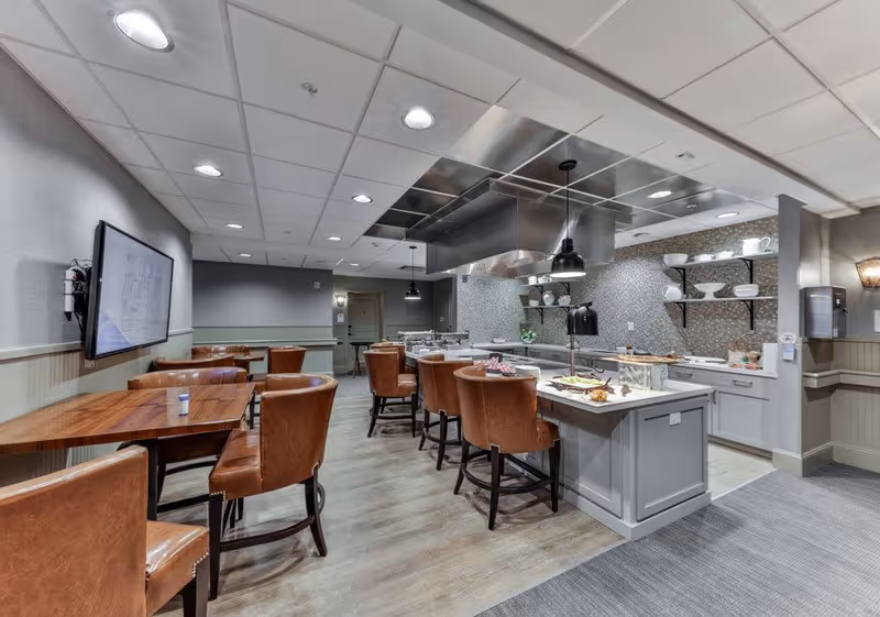 Interior view of a modern kitchen and dining area with brown leather chairs around wooden tables and a kitchen island with food trays. The kitchen has open shelves with white dishes, a large stainless steel range hood, and pendant lights hanging from the ceiling.