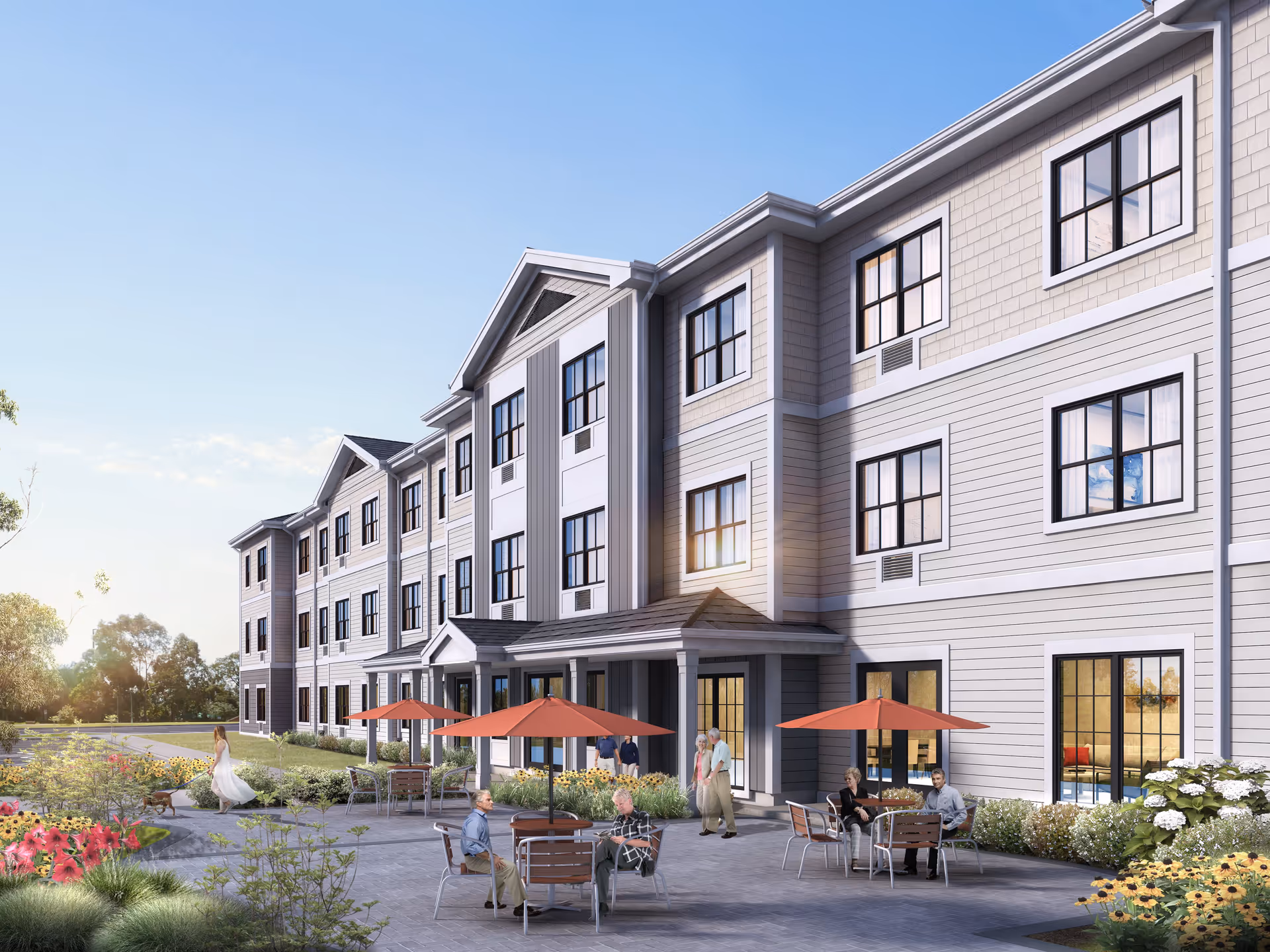 Exterior view of a three-story light-gray senior living building with an outdoor patio area, tables with red umbrellas, landscaped gardens, and people sitting and walking.