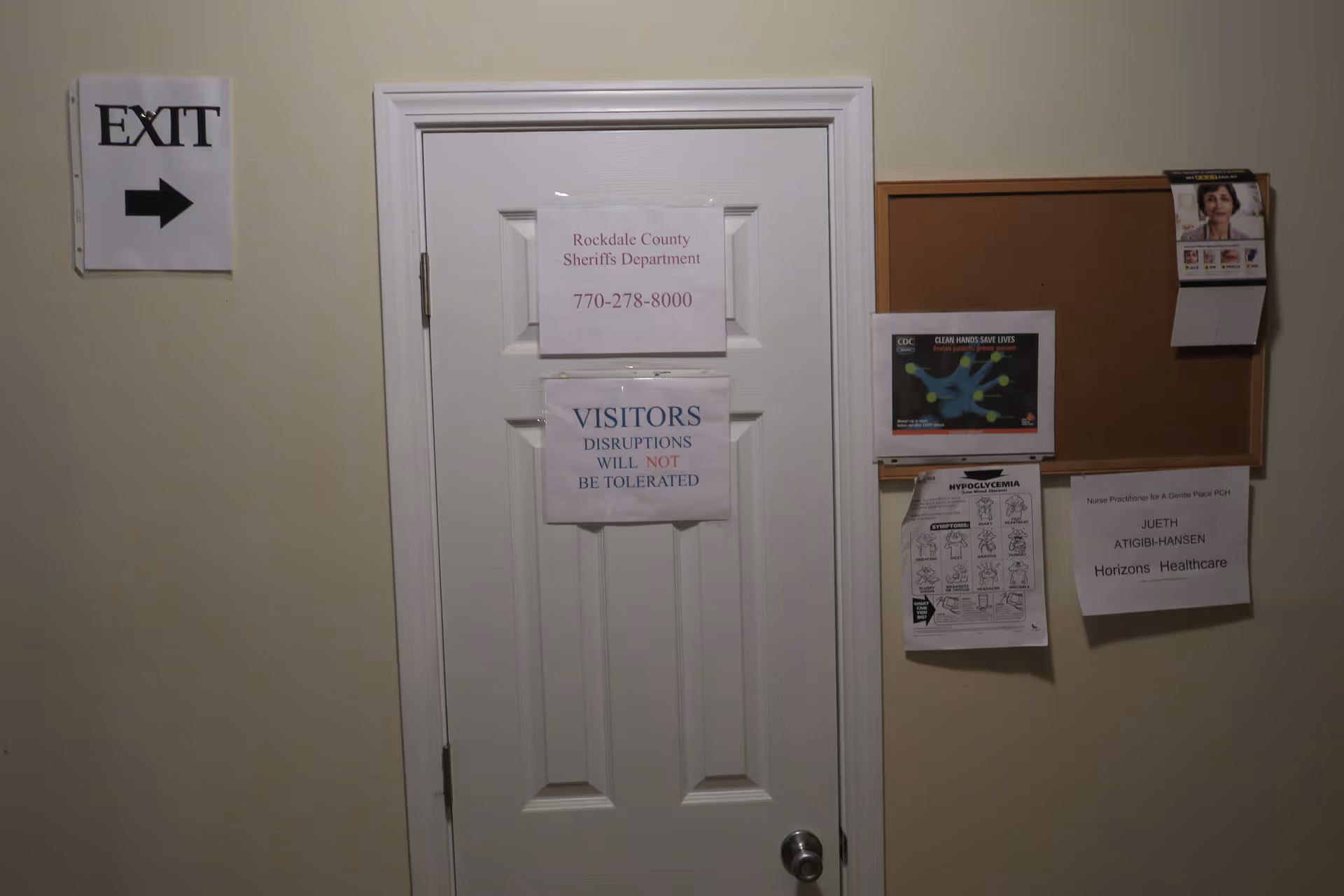 White interior door with posted notices, an 'EXIT' sign to the left, and a bulletin board with flyers on the right.