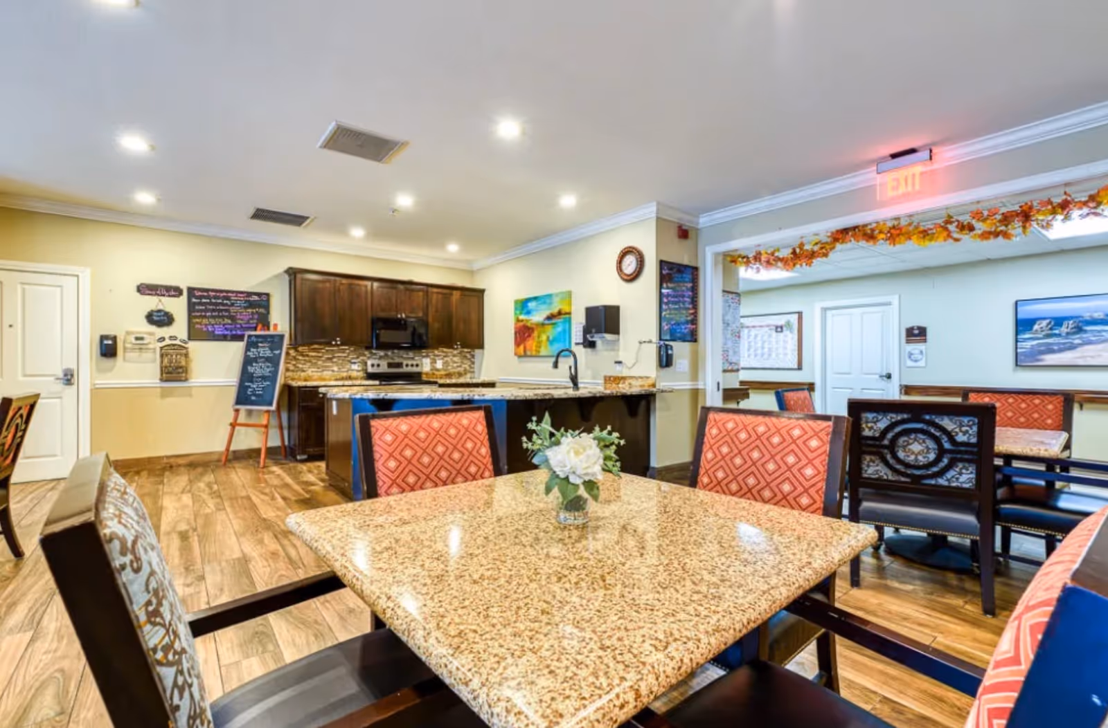 Interior view of a dining area in McMinnville Memory Care facility featuring a granite table with a small flower arrangement, surrounded by chairs with patterned upholstery. In the background, there is a kitchen area with dark wooden cabinets, a stove, microwave, and a sink. The room has wooden flooring, recessed ceiling lights, and walls decorated with colorful paintings and menu boards. An exit sign and autumn-themed garland are visible above a doorway leading to another room with additional seating.