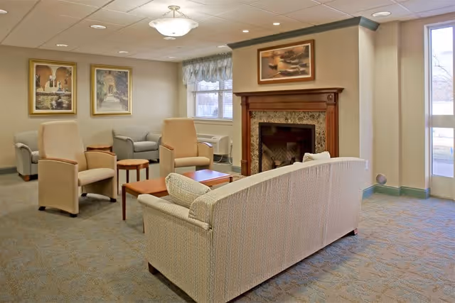 A cozy living room area in a senior living facility with a patterned sofa, two beige armchairs, a wooden coffee table, and a fireplace with a wooden mantle. The room has carpeted floors, framed artwork on the walls, a window with a valance, and a glass door letting in natural light.