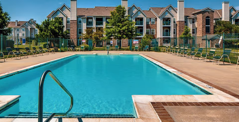 Outdoor swimming pool with clear blue water surrounded by a concrete deck and multiple green metal chairs. In the background, there are multi-story residential buildings with balconies and trees.