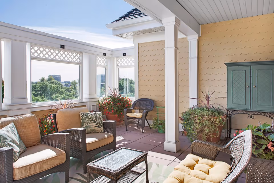 A bright and airy outdoor patio area with comfortable wicker seating including cushioned chairs and a loveseat, a glass-top coffee table, potted plants, and a green cabinet against a yellow wall with decorative paneling. The patio is partially enclosed with white columns and latticework, overlooking trees and distant buildings under a clear sky.