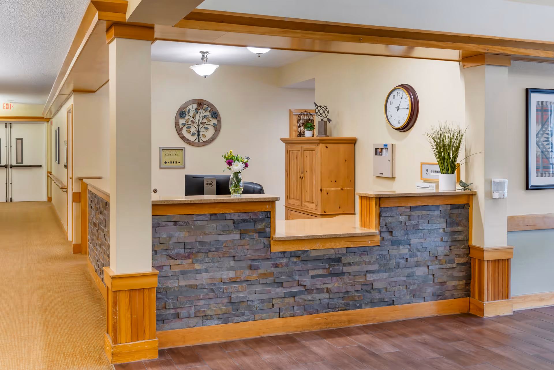 Reception desk area in a senior living facility with a stone and wood counter, a computer monitor, a vase with flowers, a wooden cabinet, a wall clock, and decorative wall art. The hallway with double doors and exit sign is visible to the left.