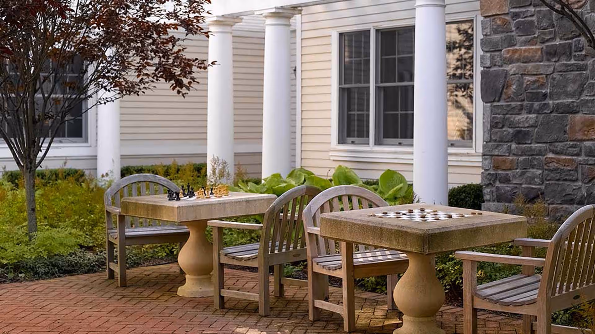 Outdoor seating area with two stone game tables featuring chess and checkers boards, surrounded by wooden chairs. The area is paved with bricks and bordered by greenery, with a building featuring white columns and stone walls in the background.