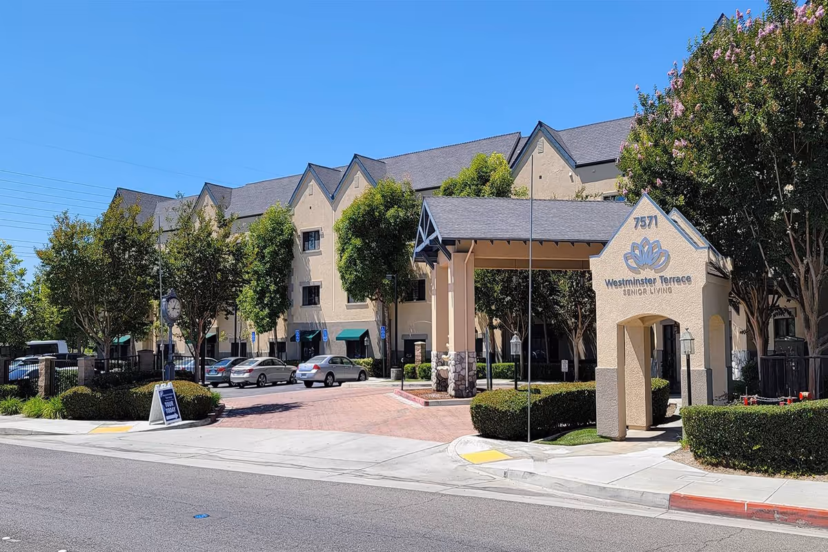 Front entrance and driveway of the Westminster Terrace senior living building with a porte-cochere, landscaped grounds, and parked cars.
