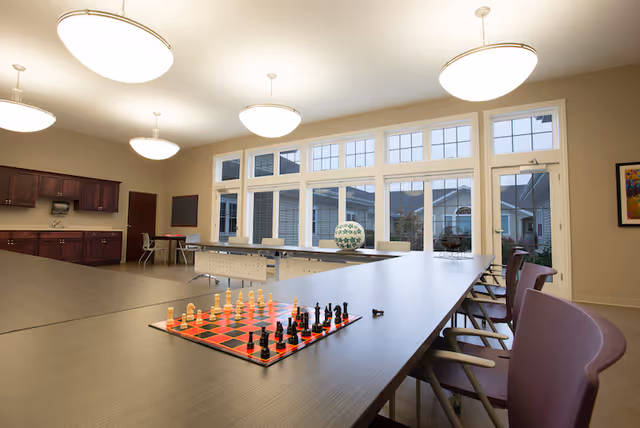 A spacious, well-lit common area with large windows showing an outdoor view. The room features a long table with a chessboard set up, several chairs, and modern ceiling lights. There are cabinets and a door along one wall, and a framed picture on another wall.