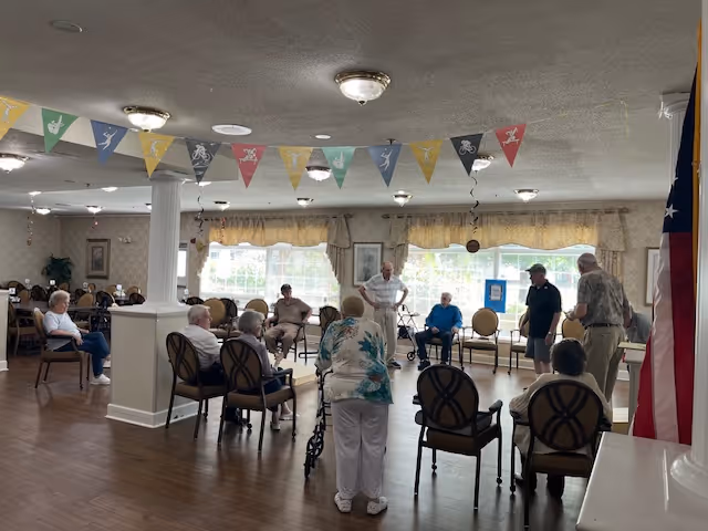 A group of elderly people seated in a spacious room with wooden floors and large windows covered with light curtains. Some individuals are sitting on chairs arranged in a circle, while others stand or use walkers. The ceiling has several light fixtures and colorful triangular pennant banners hanging across the room. An American flag is visible on the right side of the image.