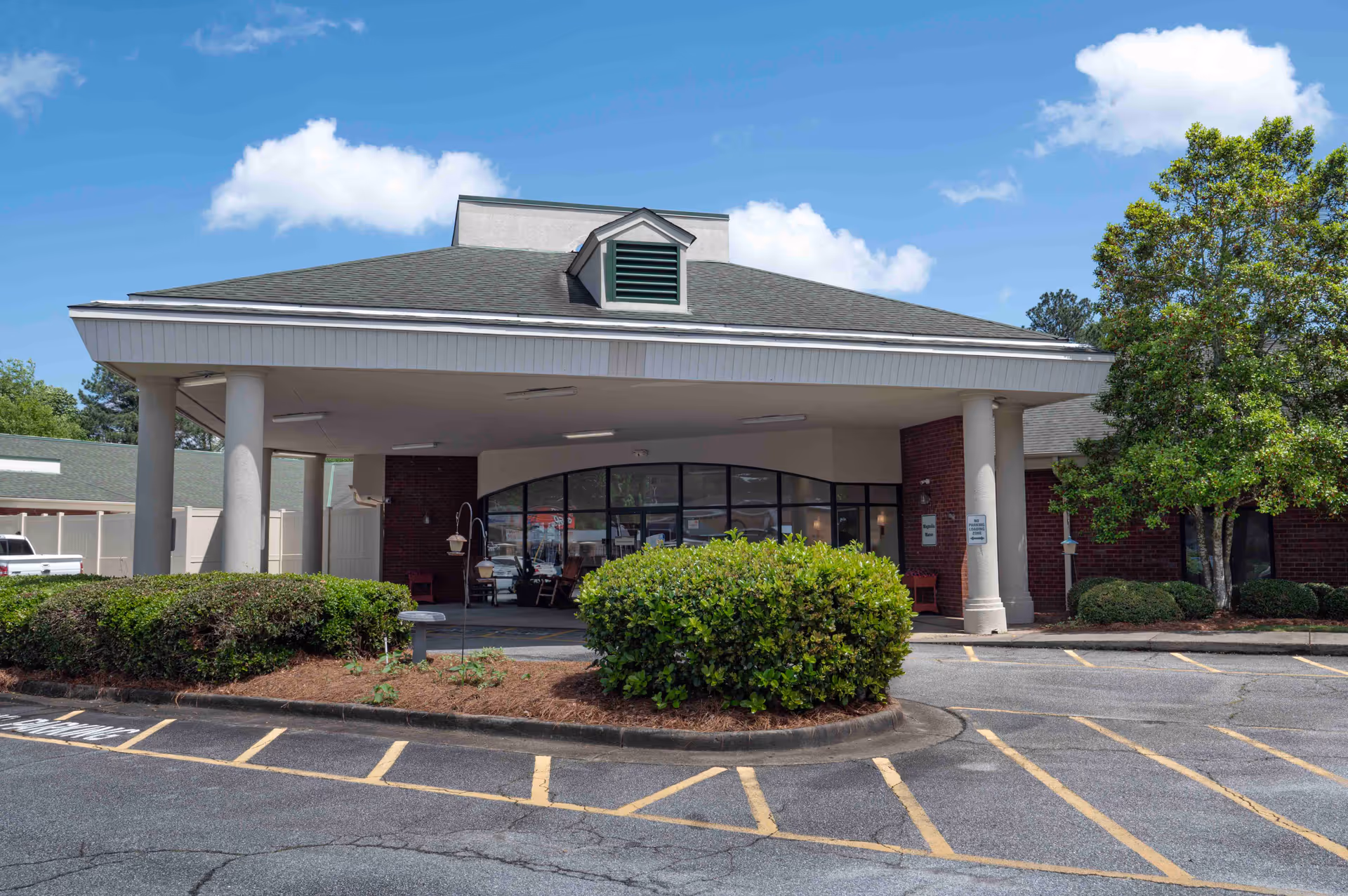 Covered porte-cochère entrance of a low-rise brick assisted living building with parking spaces and shrubs in front.