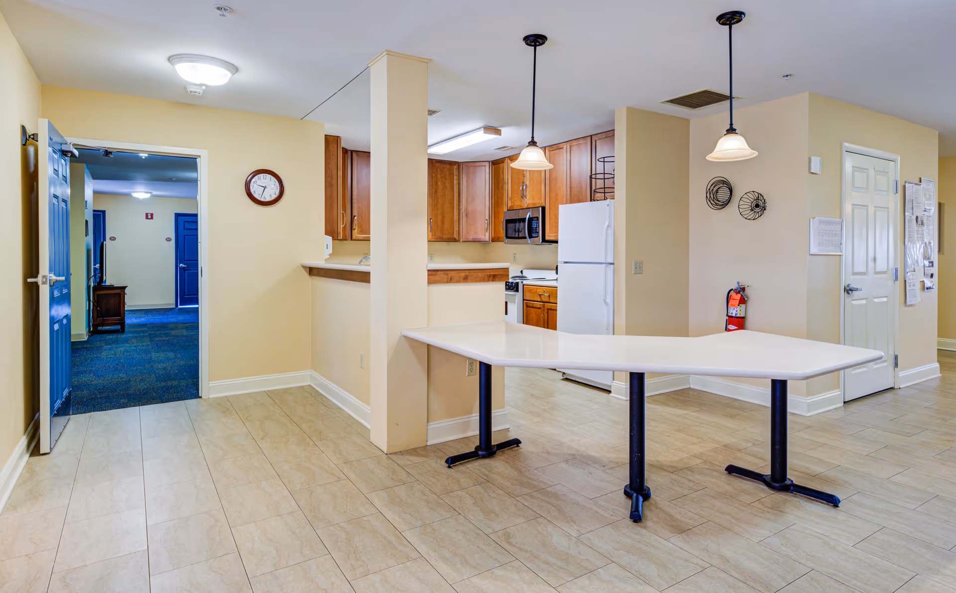Interior view of a senior living facility kitchen area with wooden cabinets, a white refrigerator, microwave, and stove. There is a large white table with black legs in the foreground, beige walls, tiled floor, two hanging pendant lights, a fire extinguisher mounted on the wall, and a clock above an open doorway leading to a carpeted hallway with blue doors.