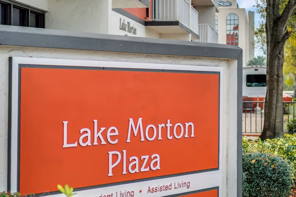 Outdoor sign for Lake Morton Plaza, a senior living facility, with part of the building and a tree visible in the background.
