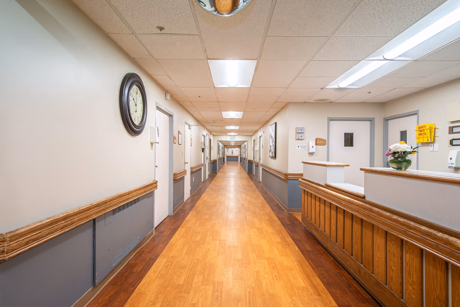 A long, well-lit hallway in a senior living facility with wooden flooring and beige walls. On the right side, there is a reception desk with a flower vase on it. Several closed doors line both sides of the hallway, and a large clock is mounted on the left wall. Fluorescent ceiling lights illuminate the corridor.