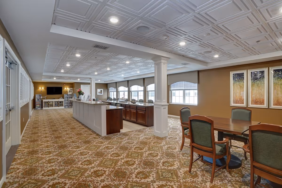 Spacious senior living common area with patterned carpet, a long counter with granite top, several chairs around a wooden table, large windows with blinds, and framed artwork on the walls. The ceiling has decorative panels with recessed lighting.