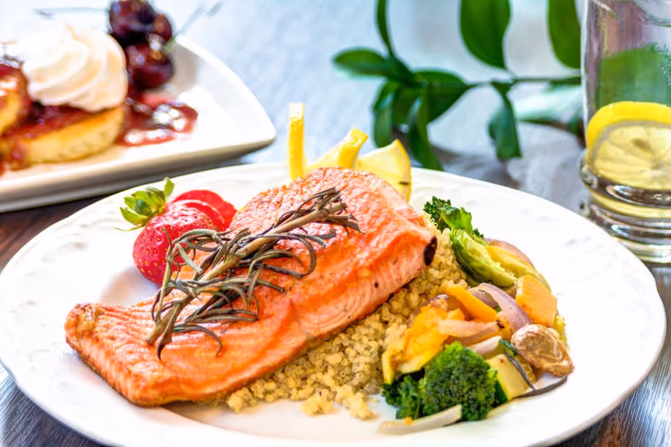 A plated meal featuring a cooked salmon fillet garnished with herbs, served on a bed of quinoa with a side of mixed steamed vegetables including broccoli, carrots, and onions. The plate also includes two strawberries and lemon wedges. In the background, there is a dessert plate with a pastry topped with whipped cream and berries, and a glass of water with lemon slices.