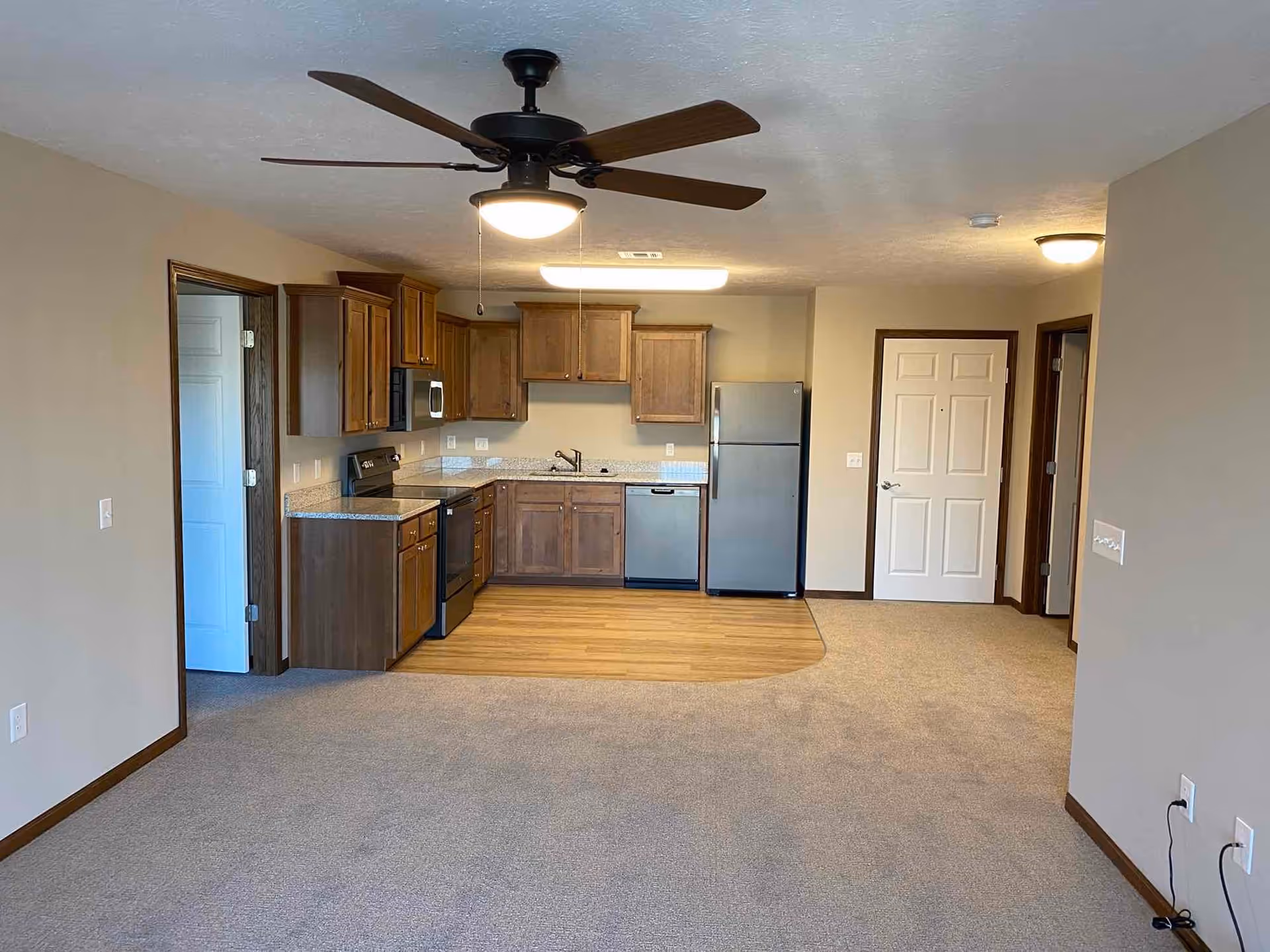 An empty apartment interior featuring a kitchen area with wooden cabinets, stainless steel appliances including a refrigerator, stove, microwave, and dishwasher. The kitchen floor is wood, transitioning to carpet in the living area. There is a ceiling fan with a light fixture above, beige walls, and two doors visible in the background.