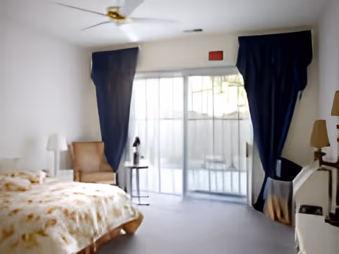 Sunlit bedroom with a bed, armchair, ceiling fan, sliding glass door with dark curtains, and small side tables.