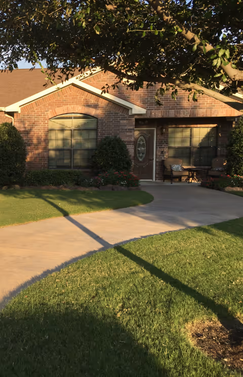 Front exterior view of a single-story brick house with a brown shingled roof, a decorative glass front door, two large windows with blinds, a concrete walkway leading to the entrance, green lawn, bushes, flowers, and a large tree casting shadows over the scene.