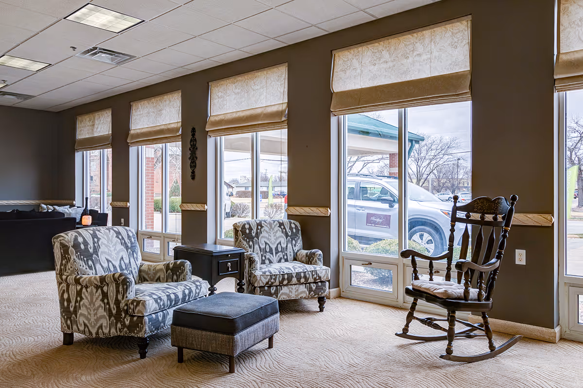 A cozy sitting area in a senior living facility with two patterned armchairs, a small black side table, a cushioned ottoman, and a wooden rocking chair. Large windows with beige Roman shades let in natural light and offer a view of the outside parking area and trees.