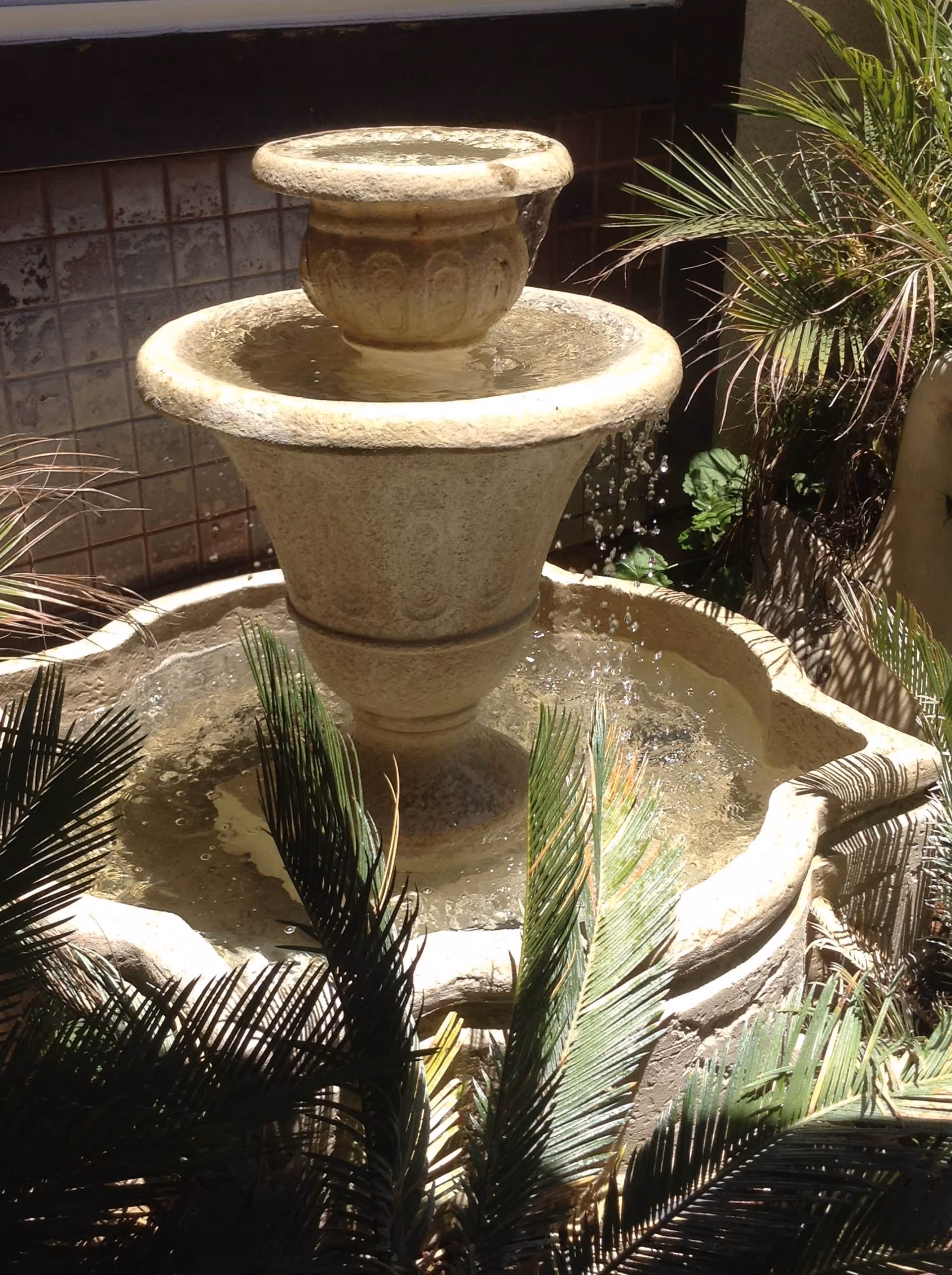 A stone water fountain with water flowing from the top tier into a larger basin below, surrounded by green palm leaves and plants in an outdoor setting.