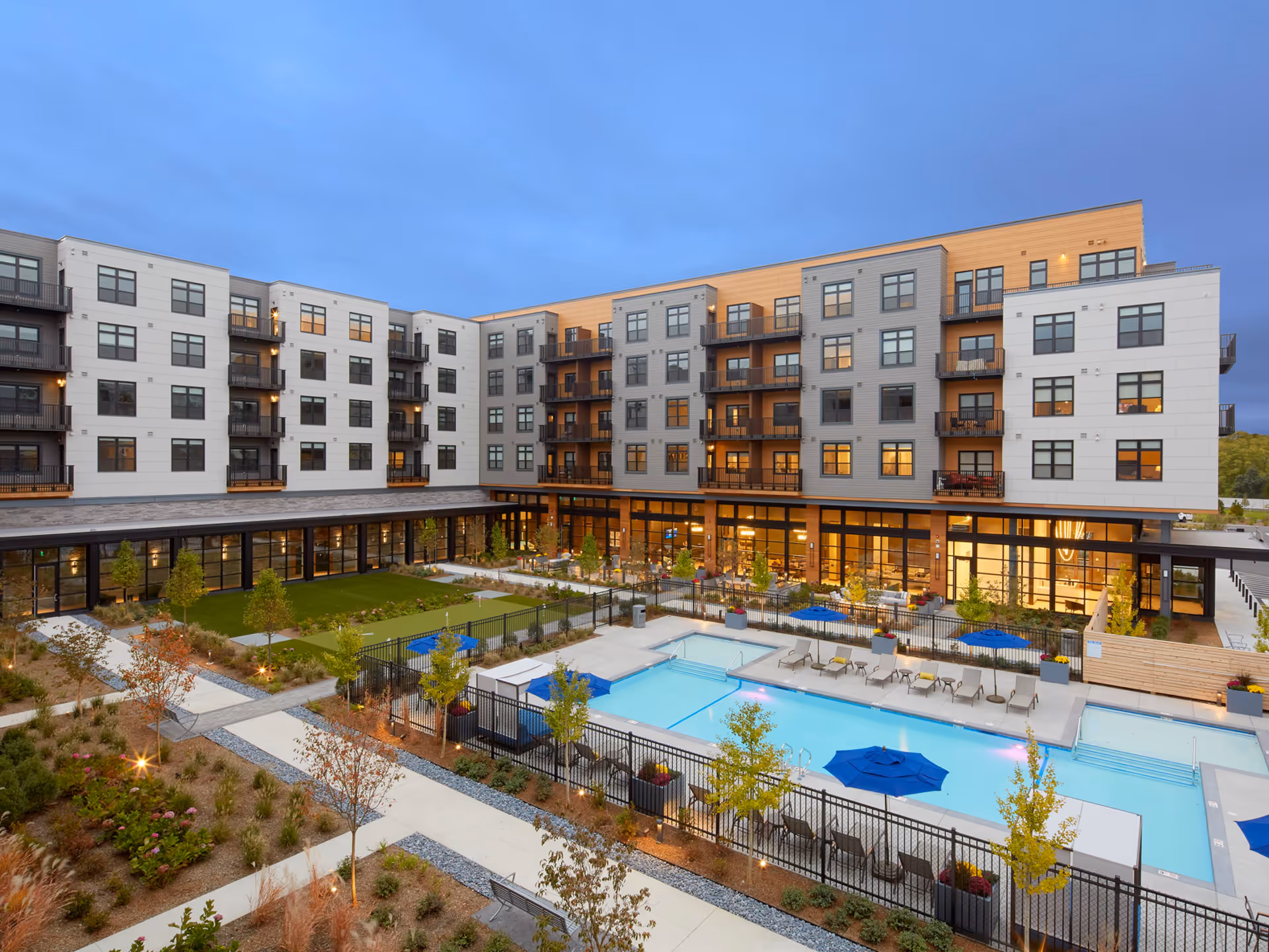 View of a modern senior living facility courtyard at dusk with a swimming pool surrounded by lounge chairs and blue umbrellas, landscaped garden areas, and a multi-story building with balconies and large windows.