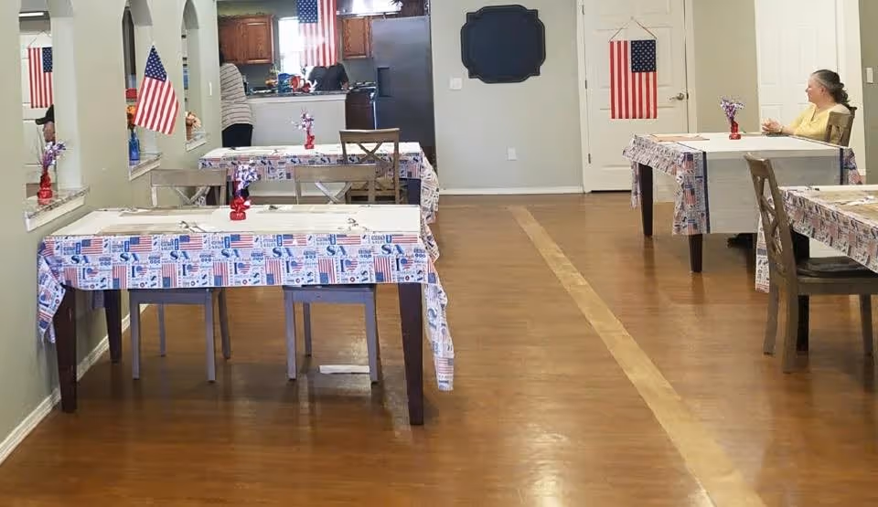 Dining room with tables covered in patriotic tablecloths, chairs, American flags, and a resident seated at a table.